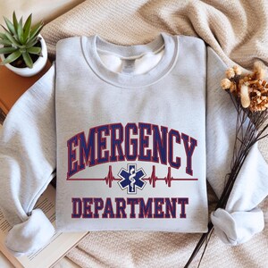 May include: A light gray sweatshirt with the words "EMERGENCY DEPARTMENT" in red and blue lettering. A medical symbol and heartbeat graphic are centered below the word "EMERGENCY". The sweatshirt is folded.