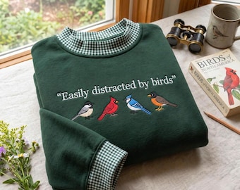 May include: A dark green sweatshirt featuring a green and white gingham collar and cuffs. The chest displays the embroidered text "Easily distracted by birds" above four colorful bird illustrations: a chickadee, cardinal, blue jay, and robin.