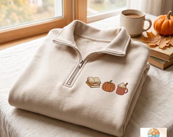Puede incluir: Una sudadera beige con cremallera corta y un pequeño gráfico bordado en el pecho izquierdo. El diseño muestra una pila de tres libros abiertos, una calabaza naranja redonda y una taza marrón con una bebida cubierta de crema batida.