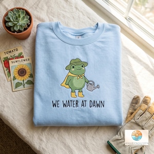 May include: A light blue crewneck sweatshirt featuring an embroidered green frog wearing a hat, yellow cape, and yellow boots, holding a small watering can. Below the frog, the text reads WE WATER AT DAWN in dark blue capital letters.