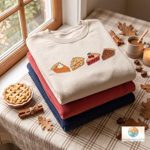 May include: A stack of three crewneck sweatshirts in cream, red, and navy blue. The top cream sweatshirt features an embroidered graphic of four pie slices: pumpkin with whipped cream, apple, cherry, and pecan, arranged in a horizontal row.