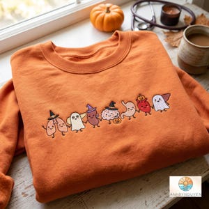 May include: An orange crewneck sweatshirt featuring an embroidered line of cartoon human organs and ghosts holding hands. The characters include lungs, a ghost, a kidney, a brain, a stomach, a heart, and a ghost, all wearing small witch hats.