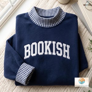 May include: A navy blue sweatshirt featuring the word BOOKISH in white block letters across the chest. The garment has a mock neck collar and sleeve cuffs lined with a navy and white gingham check pattern. A small logo with a sunset icon is visible.