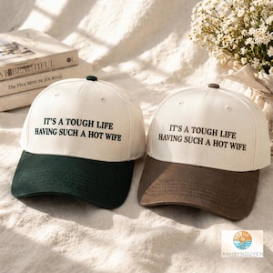 Puede incluir: Dos gorras de b&eacute;isbol con coronas color blanco roto y viseras oscuras. La gorra izquierda tiene una visera verde oscuro y la derecha una marr&oacute;n oscuro. Ambas presentan el texto negro bordado: IT'S A TOUGH LIFE HAVING SUCH A HOT WIFE.