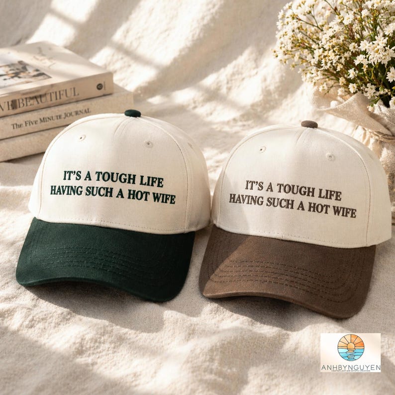 Puede incluir: Dos gorras de b&eacute;isbol con coronas color blanco roto y viseras oscuras. La gorra izquierda tiene una visera verde oscuro y la derecha una marr&oacute;n oscuro. Ambas presentan el texto negro bordado: IT'S A TOUGH LIFE HAVING SUCH A HOT WIFE.