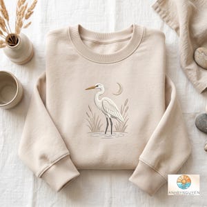 May include: A beige crewneck sweatshirt featuring an embroidered graphic of a white heron standing in water among reeds with a crescent moon above. The design is centered on the chest, and the garment has long sleeves with ribbed cuffs and a ribbed neckline.
