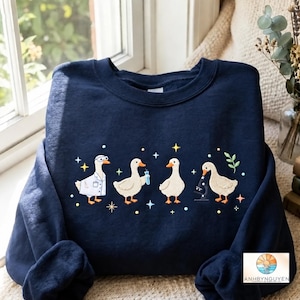 May include: A navy blue crewneck sweatshirt featuring an embroidered graphic of four white ducks engaged in scientific activities. The ducks are depicted wearing a lab coat with goggles, holding a blue test tube, and using a black microscope.
