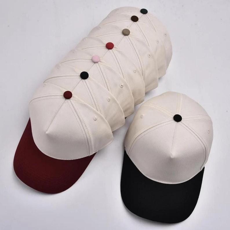 Puede incluir: Una pila de gorras de b&eacute;isbol con coronas de color blanco roto y viseras de varios colores, incluyendo granate y negro. Cada gorra presenta un bot&oacute;n a juego en el centro superior de la corona. Las gorras est&aacute;n dispuestas en una fila vertical.