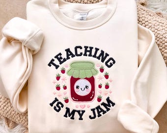 May include: Cream-colored sweatshirt with the embroidered phrase "TEACHING IS MY JAM." The design features a smiling jar of jam with a green lid, surrounded by strawberries. The sweatshirt has long sleeves and a crew neck.