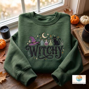 Puede incluir: Una sudadera de cuello redondo en color verde oliva oscuro con un diseño bordado que incluye la palabra WITCHY en letra cursiva negra. El gráfico muestra un sombrero de bruja morado, tres frascos de pociones, una luna creciente y un gato.