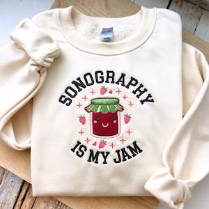 Peut inclure: Sweat-shirt crème avec la phrase brodée "SONOGRAPHY IS MY JAM". Le motif présente un pot de confiture souriant avec des fraises et des accents en forme de croix. Les manches sont resserrées.