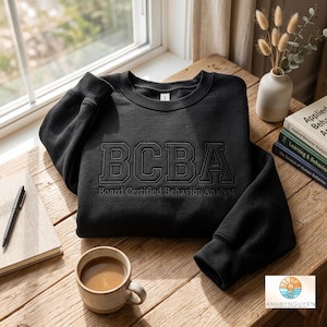 May include: A black crewneck sweatshirt featuring the embroidered letters BCBA in a collegiate font, with the text Board Certified Behavior Analyst stitched directly underneath in a smaller, matching black thread on the chest area.