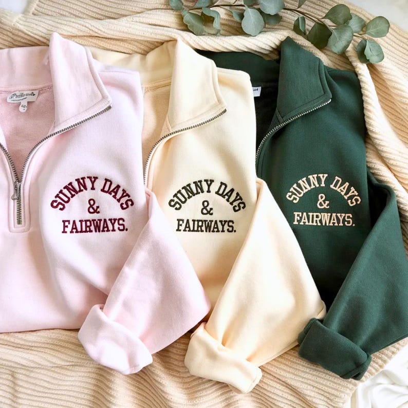 Puede incluir: Cuatro sudaderas con cremallera de un cuarto en rosa pastel, crema y verde oscuro. Cada sudadera tiene el texto bordado "SUNNY DAYS & FAIRWAYS" en un color contrastante. Las sudaderas est&aacute;n dobladas sobre una manta color crema.