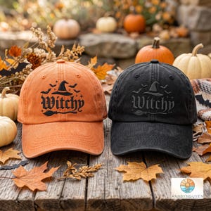 Könnte beinhalten: Eine orangefarbene Baseballkappe und eine schwarze Baseballkappe, beide mit einer Stickerei eines Hexenhuts, eines Besens, Fledermäusen und dem Wort Witchy in gotischer Schrift, jeweils mit gebogenem Schirm und Ösen versehen.