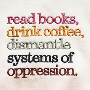 May include: Embroidered text on a cream-colored fabric reads "read books, drink coffee, dismantle systems of oppression." The text is in various colors: pink, gold, silver, black, and brown. The embroidery has a textured appearance.