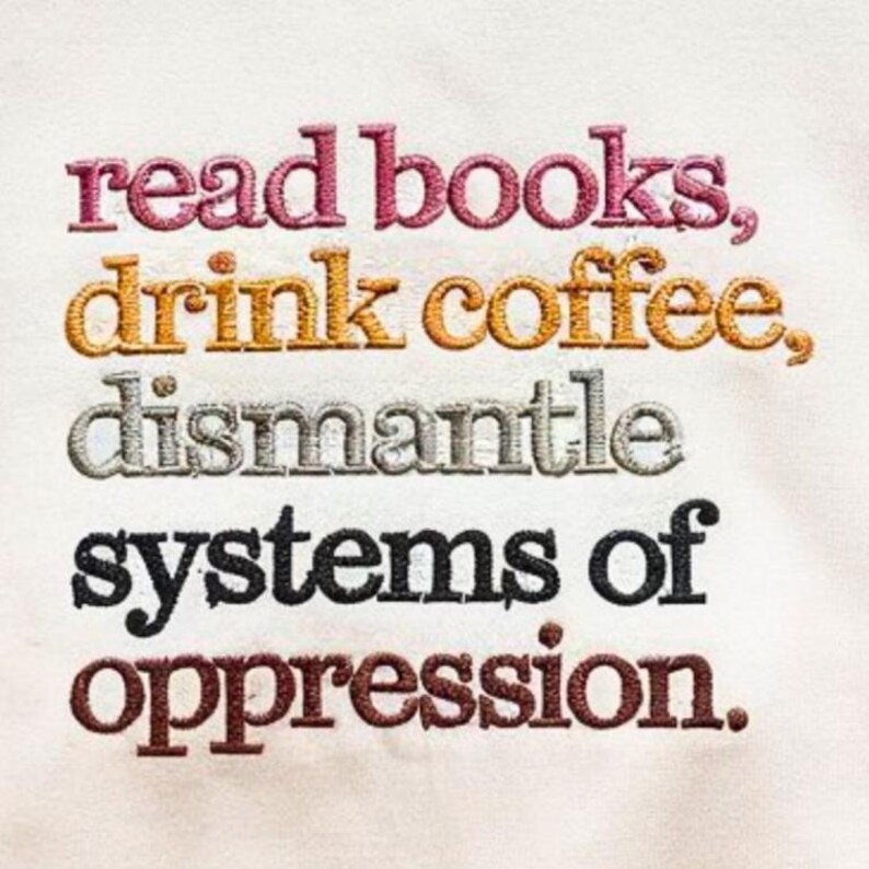 May include: Embroidered text on a cream-colored fabric reads "read books, drink coffee, dismantle systems of oppression." The text is in various colors: pink, gold, silver, black, and brown. The embroidery has a textured appearance.