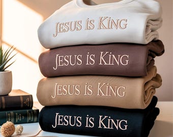 May include: A stack of four sweatshirts in cream, brown, tan, and black. Each sweatshirt has the embroidered text "JESUS is KING" in a complementary color. The sweatshirts are neatly folded and stacked on a white surface.