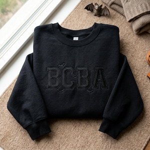May include: A black crewneck sweatshirt featuring the letters BCBA embroidered in a matching black thread on the chest. The design includes subtle embroidered details of small bats and spiderwebs surrounding the bold, block-style lettering.