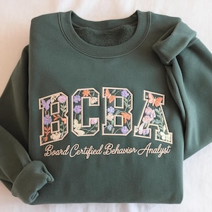May include: Dark green sweatshirt featuring the embroidered letters "BCBA" filled with colourful flowers and butterflies. The text "Board Certified Behavior Analyst" is embroidered below. The sweatshirt is folded on a white surface.