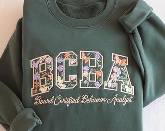 May include: Dark green sweatshirt featuring the embroidered letters "BCBA" filled with colourful flowers and butterflies. The text "Board Certified Behavior Analyst" is embroidered below. The sweatshirt is folded on a white surface.
