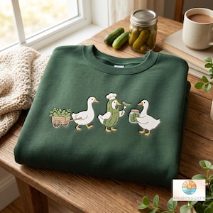 Op de afbeelding: Een donkergroene sweater met ronde hals en een geborduurde afbeelding van drie witte ganzen. Eén gans trekt een houten kar vol augurken, een andere draagt een koksmuts en augurkenkostuum met een vork, en de derde houdt een glazen pot vast.