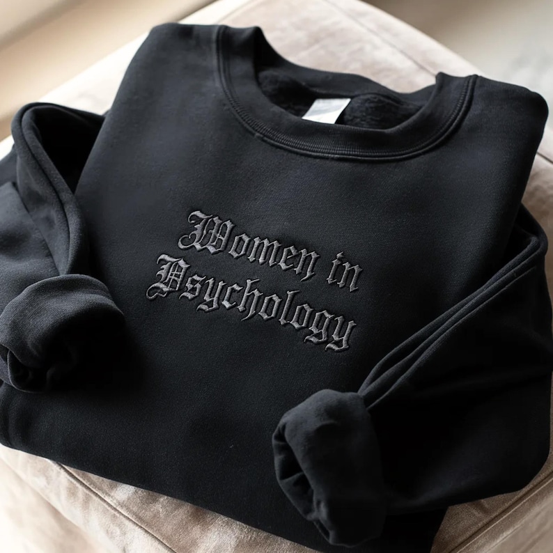 May include: Black sweatshirt featuring the embroidered phrase "Women in Psychology" in a gothic font. The crew neck sweatshirt has long sleeves and is made of a soft material. The item is folded on a neutral-toned surface.