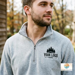 May include: A light grey quarter-zip fleece pullover featuring a black embroidered graphic on the left chest. The design displays a cluster of pine trees above the text "YOUR LOGO HERE" with a horizontal arrow pointing in both directions.