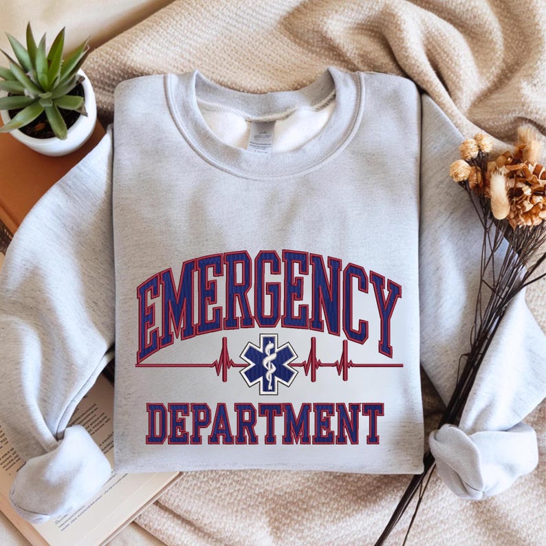 May include: A light gray sweatshirt with the words "EMERGENCY DEPARTMENT" in red and blue lettering. A medical symbol and heartbeat graphic are centered below the word "EMERGENCY". The sweatshirt is folded.