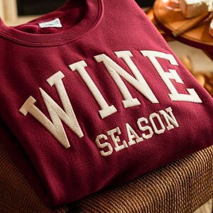 May include: A burgundy sweatshirt with the words "WINE SEASON" embroidered in a cream-colored, collegiate-style font. The sweatshirt is folded and resting on a brown surface.