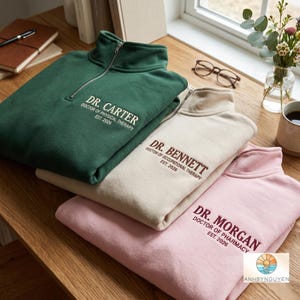Puede incluir: Tres sudaderas con cremallera de un cuarto en verde oscuro, crema y rosa claro. Cada una presenta texto bordado: DR. CARTER DOCTOR OF PHYSICAL THERAPY EST. 2026, DR. BENNETT DOCTOR OF OCCUPATIONAL THERAPY EST. 2026 y DR. MORGAN DOCTOR OF PHARMACY EST. 2026.