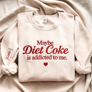 Może przedstawiać: Kremowa bluza z haftowanym napisem "Maybe Diet Coke is addicted to me" czerwoną czcionką. Małe czerwone serce znajduje się pod tekstem, a puszka Diet Coke jest wyhaftowana na lewym rękawie.