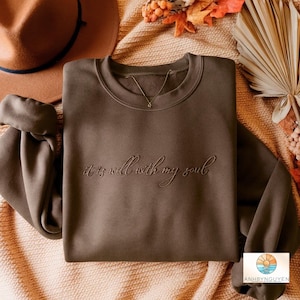 Puede incluir: Una sudadera de cuello redondo en color marrón oscuro con el texto bordado "it is well with my soul" en una fuente cursiva de color marrón oscuro a juego en el centro del pecho. La prenda tiene mangas largas y un cuello acanalado.