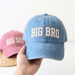 以下が含まれることがあります： デニム素材の野球帽。水色の帽子には白い文字で「BIG BRO」、ピンクの帽子には「BIG SIS」と書かれています。どちらの帽子もカーブしたつばと調節可能なストラップが付いています。