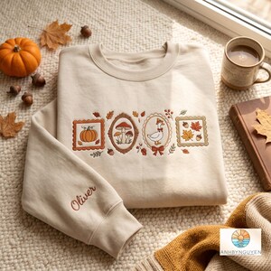 Peut inclure: Un sweat-shirt beige à col rond orné de broderies sur le thème de l'automne, représentant une citrouille, des champignons, une oie portant un chapeau et des feuilles d'érable dans des cadres. Le prénom Oliver est brodé en lettres cursives marron sur le poignet gauche.