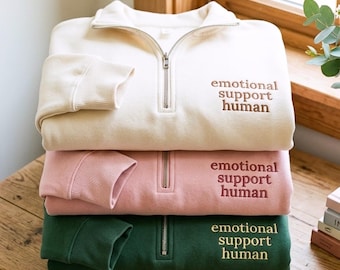May include: Three quarter-zip sweatshirts in cream, pink, and green are stacked. Each sweatshirt features the embroidered text "emotional support human". The sweatshirts are neatly folded on a wooden surface.