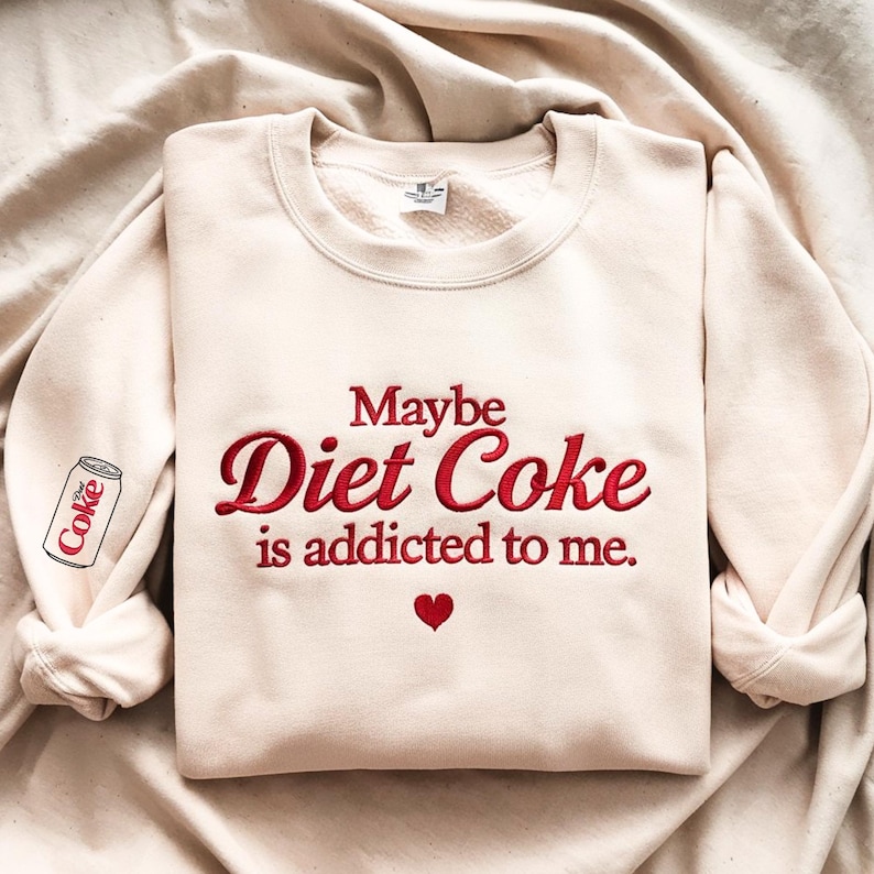 Może przedstawiać: Kremowa bluza z haftowanym napisem "Maybe Diet Coke is addicted to me" czerwoną czcionką. Małe czerwone serce znajduje się pod tekstem, a puszka Diet Coke jest wyhaftowana na lewym rękawie.