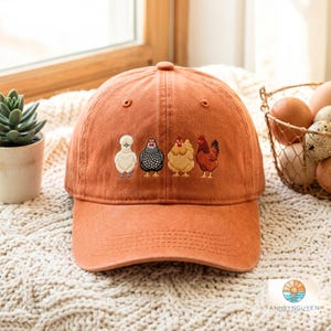 May include: A rust-coloured baseball cap featuring an embroidered design of four distinct chickens in a row. The embroidery depicts a white fluffy chicken, a black and white speckled chicken, a light brown chicken, and a dark red chicken.