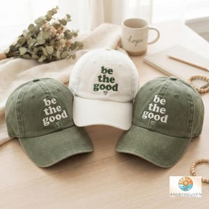 Könnte beinhalten: Drei Baseballkappen, eine weiße und zwei olivgrüne, jeweils mit dem gestickten Text be the good und einem kleinen Herzsymbol. Die weiße Kappe hat dunkelgrüne Schrift, während die beiden olivgrünen Kappen weiße Schrift haben.