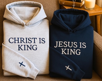 May include: Two hooded sweatshirts, one light gray and one navy blue, are displayed. The light gray sweatshirt has the text "CHRIST IS KING" and a cross on the sleeve. The navy blue sweatshirt has the text "JESUS IS KING" and a cross on the sleeve.