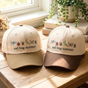 May include: Two baseball caps with a beige crown and a contrasting brim. The caps feature embroidered insects and the text "sad bug summer". One cap has a tan brim, the other a brown brim. The caps are on a wooden surface.