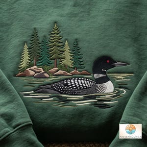 Puede incluir: Una sudadera verde oscuro con un dise&ntilde;o bordado de un colimbo nadando en el agua frente a un grupo de pinos y rocas. El ave tiene plumas con patrones en blanco y negro y un ojo rojo. El texto ANHBYNGUYEN aparece en la parte inferior.
