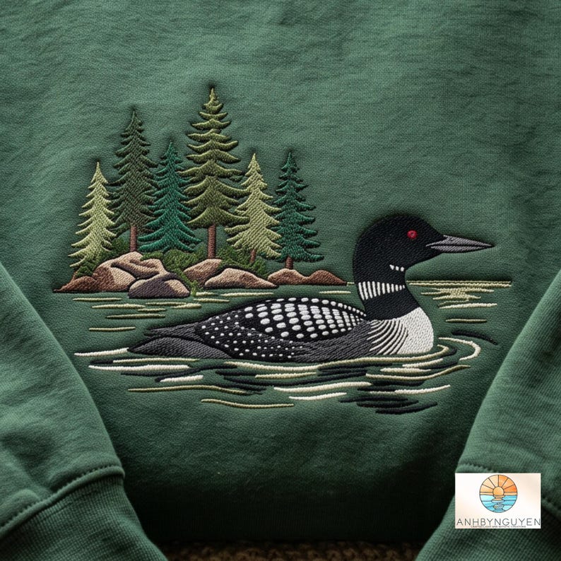 Puede incluir: Una sudadera verde oscuro con un dise&ntilde;o bordado de un colimbo nadando en el agua frente a un grupo de pinos y rocas. El ave tiene plumas con patrones en blanco y negro y un ojo rojo. El texto ANHBYNGUYEN aparece en la parte inferior.