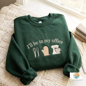 May include: A dark green crewneck sweatshirt featuring white embroidered text that reads I'll be in my office. Below the text are embroidered icons of two whisks, a tan oven mitt, and a white stand mixer with a bowl, centered on the chest.