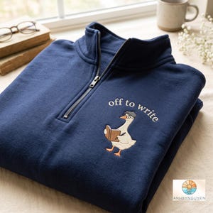 May include: A navy blue quarter-zip sweatshirt featuring an embroidered graphic of a white goose wearing a grey cap and glasses, holding a brown book and a pen. The text "off to write" is embroidered in white above the goose illustration.