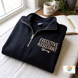 Puede incluir: Una sudadera negra con cremallera de un cuarto de largo que presenta el texto "EXECUTIVE ASSISTANT" bordado en color beige claro sobre un pequeño icono de llave a juego. La prenda tiene un tirador de cremallera metálico plateado y cuello alto.