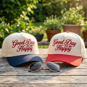 May include: Two baseball caps with the text "Good Day to be Happy" in red script. One cap has a navy blue brim, the other a red brim. Gold-framed sunglasses rest in front of the caps on a wooden surface.