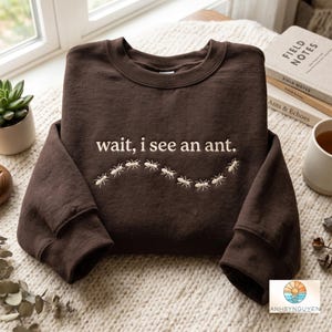 May include: A dark brown crewneck sweatshirt featuring the embroidered text "wait, i see an ant." in a light cream colour. Below the text, a line of small, embroidered cream-coloured ants follows a curved, wavy path across the centre of the chest.
