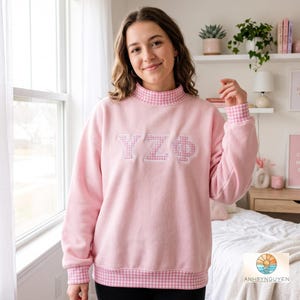 Puede incluir: Una sudadera de manga larga de color rosa claro con cuello alto, puños y dobladillo inferior con un patrón de cuadros vichy en rosa y blanco. El centro del pecho muestra las letras griegas YZΦ en tela de aplicación de cuadros vichy.