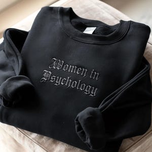 May include: Black sweatshirt featuring the embroidered phrase "Women in Psychology" in a gothic font. The crew neck sweatshirt has long sleeves and is made of a soft material. The garment is folded on a neutral-toned surface.
