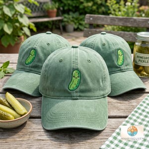 Op de afbeelding: Drie saliegroene baseballpetten van katoen, elk voorzien van een geborduurd cartoon-augurkfiguurtje op het voorpaneel. De petten hebben een gestructureerde kroon, een gebogen klep met stiksels en ventilatiegaatjes op de bovenste panelen.